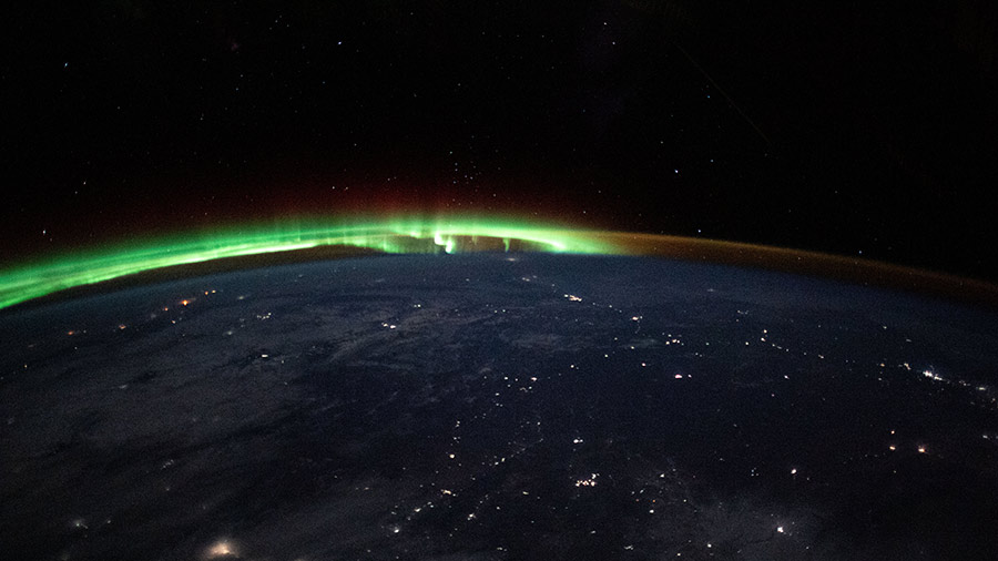 An aurora dances in the horizon of Earth’s atmosphere as city lights shine through clouds cast over Mongolia.